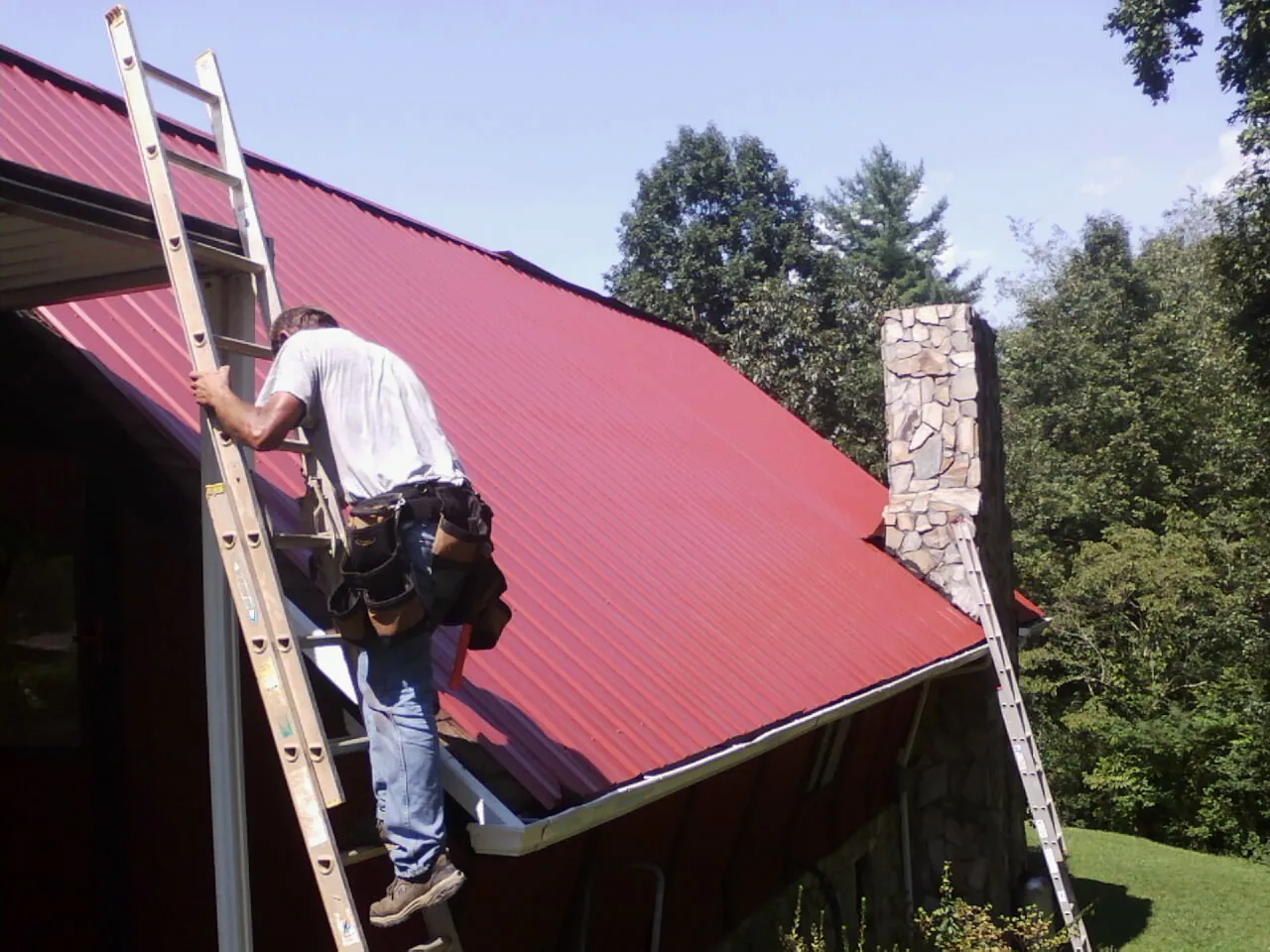 Roofer on a ladder beside a red metal roof and stone chimney — Soffit & Fascia Repair by Roofing Brothers in Martville